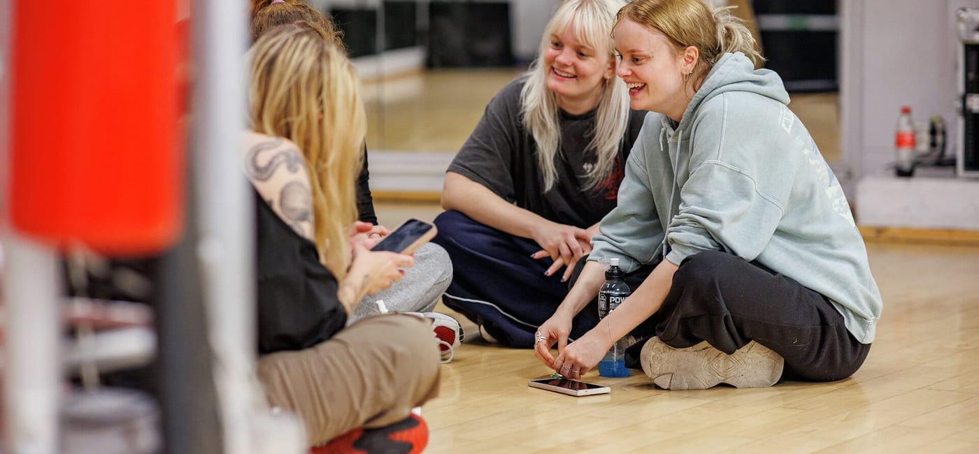 En gjeng jenter sitter på gulvet i et dansestudio. De smiler og snakker sammen.
