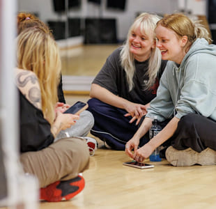 En gjeng jenter sitter på gulvet i et dansestudio. De smiler og snakker sammen.