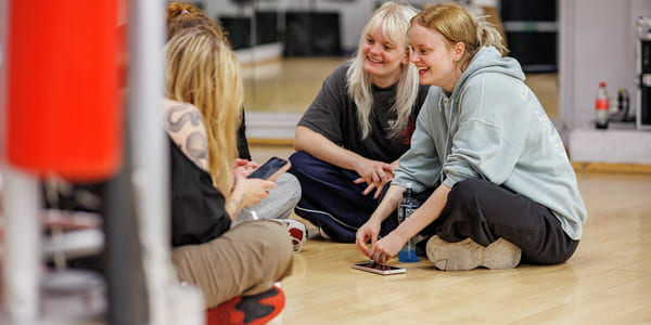 En gjeng jenter sitter på gulvet i et dansestudio. De smiler og snakker sammen.