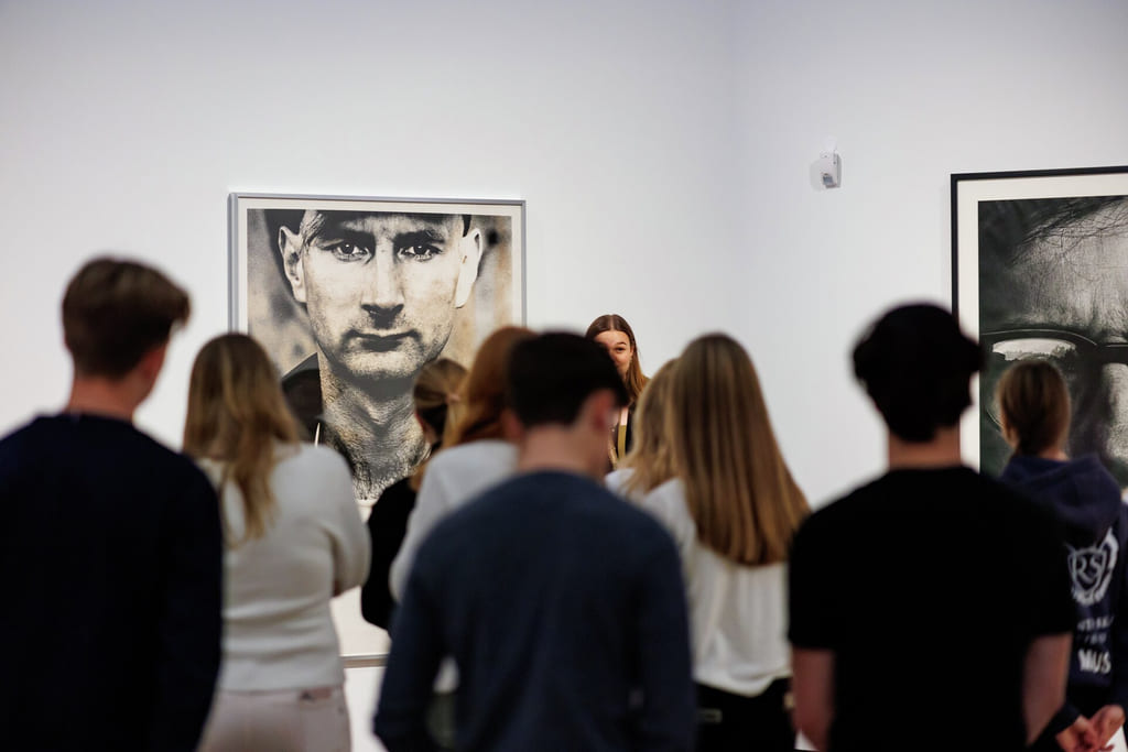 Gruppe mennesker ser på et stort portrettfotografi i et kunstgalleri, med annet innrammet kunstverk synlig på hvite vegger.