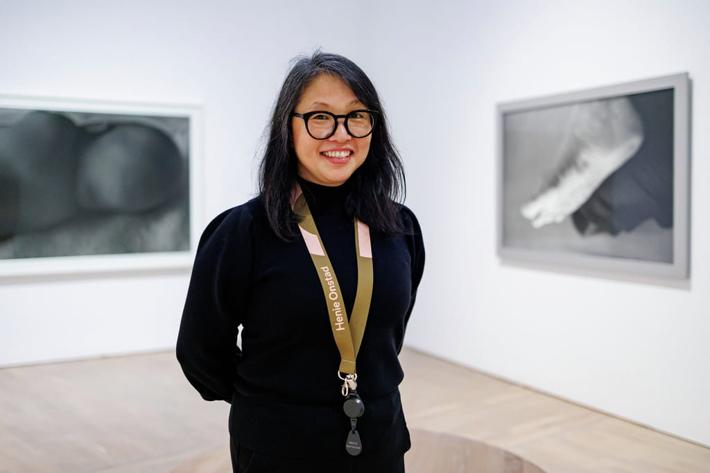 Kvinne i svart antrekk smiler i kunstgalleri, bruker briller og snor. To innrammede kunstverk synlige på hvite vegger.