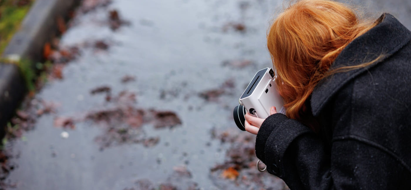 Person med rødt hår som holder kamera, fotograferer vått fortau med spredte høstblader på en dyster dag.