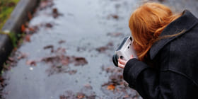 Person med rødt hår som holder kamera, fotograferer vått fortau med spredte høstblader på en dyster dag.