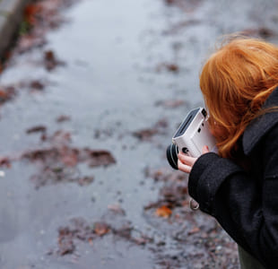 Person med rødt hår som holder kamera, fotograferer vått fortau med spredte høstblader på en dyster dag.