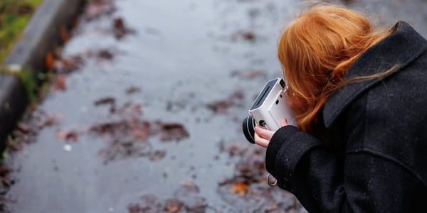Person med rødt hår som holder kamera, fotograferer vått fortau med spredte høstblader på en dyster dag.