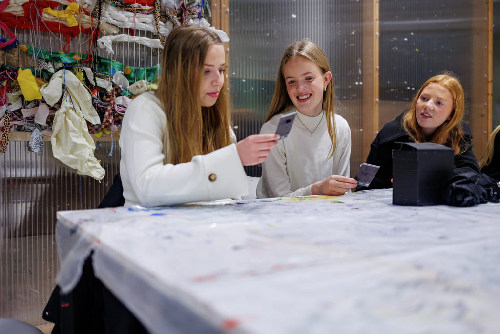 Tre unge kvinner sitter ved et bord med kort, smiler og samhandler. Fargerike stoffbiter henger i bakgrunnen.