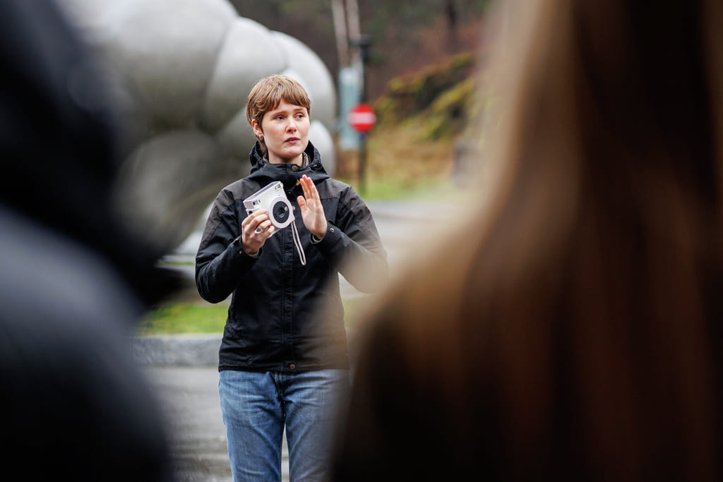 Kvinne i svart jakke holder kamera og gestikulerer med hånden. Uskarp forgrunn, utendørsmiljø med synlig stoppskilt.