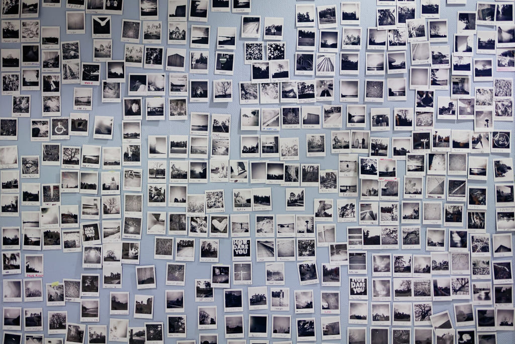 En vegg dekket med hundrevis av små svart-hvitt-polaroidfotografier, som skaper en mosaikklignende utstilling på en lyseblå bakgrunn.