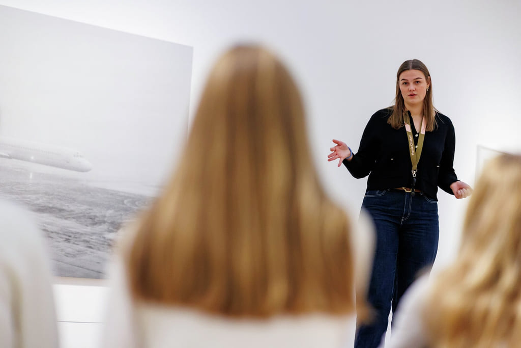 Kvinne i svart topp gestikulerer mens hun snakker til publikum i galleriet, med landskapskunst synlig på hvit vegg.