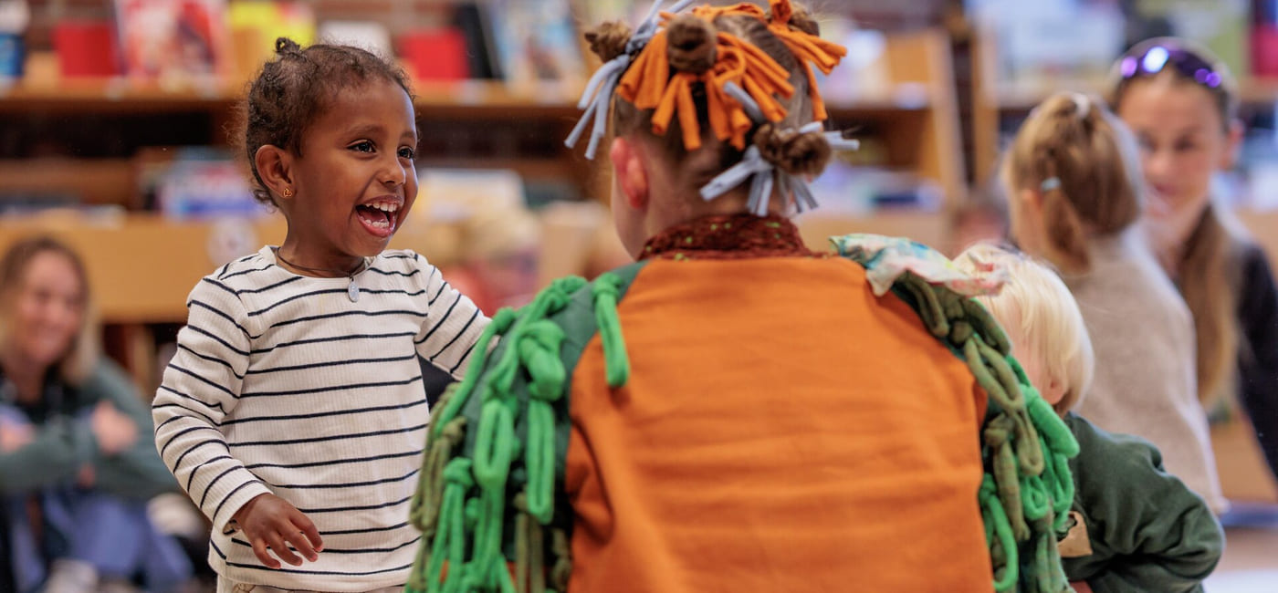 Smilende barn samhandler med utkledd artist i fargerikt antrekk på et arrangement på biblioteket. Andre barn og voksne er synlige i bakgrunnen.