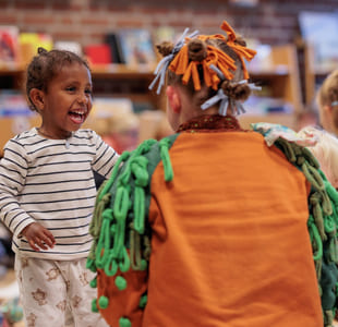 Smilende barn samhandler med utkledd artist i fargerikt antrekk på et arrangement på biblioteket. Andre barn og voksne er synlige i bakgrunnen.
