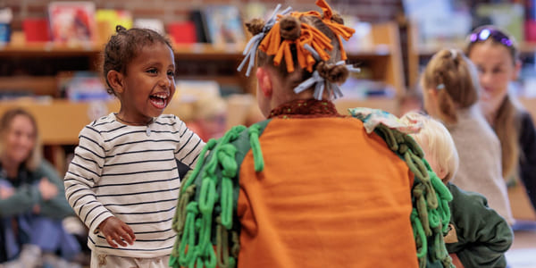 Smilende barn samhandler med utkledd artist i fargerikt antrekk på et arrangement på biblioteket. Andre barn og voksne er synlige i bakgrunnen.