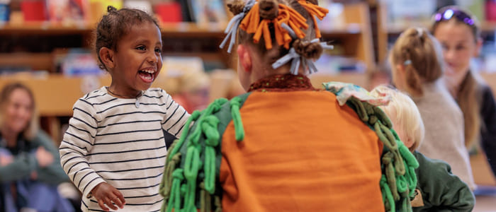 Smilende barn samhandler med utkledd artist i fargerikt antrekk på et arrangement på biblioteket. Andre barn og voksne er synlige i bakgrunnen.