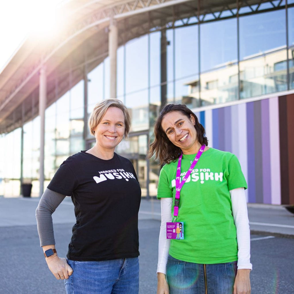 To damer ser i kamera. På t-skjortene deres står det 