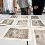 Gruppe som undersøker forskjellige skisser og etsninger lagt ut på et bord, med folks torsoer synlige i bakgrunnen.