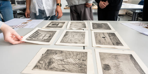 Gruppe som undersøker forskjellige skisser og etsninger lagt ut på et bord, med folks torsoer synlige i bakgrunnen.