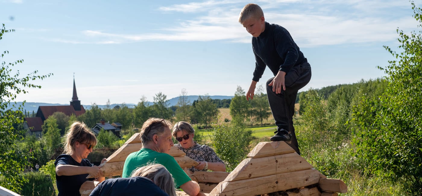 Fem personer bygger trestruktur utendørs, barn balanserer på toppen. Landlig landskap med kirkespir synlig i bakgrunnen.