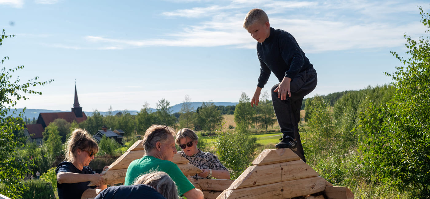 Fem personer bygger trestruktur utendørs, barn balanserer på toppen. Landlig landskap med kirkespir synlig i bakgrunnen.