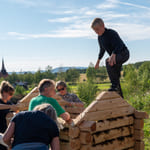 Fem personer bygger trestruktur utendørs, barn balanserer på toppen. Landlig landskap med kirkespir synlig i bakgrunnen.