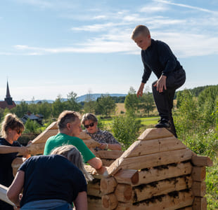 Fem personer bygger trestruktur utendørs, barn balanserer på toppen. Landlig landskap med kirkespir synlig i bakgrunnen.