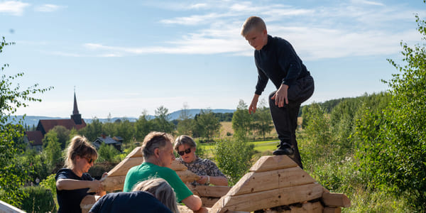 Fem personer bygger trestruktur utendørs, barn balanserer på toppen. Landlig landskap med kirkespir synlig i bakgrunnen.