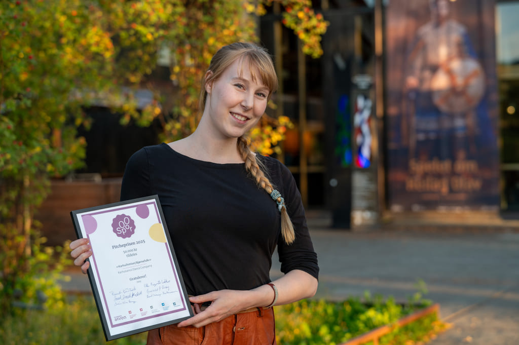 Ung kvinne med blond flette som holder sertifikat utendørs, smilende foran fargerik høstløvverk og bygningsfasade.