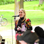 Kvinne i fargerik blomstrete jakke snakker ved mikrofon utendørs, holder en bok. Publikum er synlige i en grønn park med sykkel.