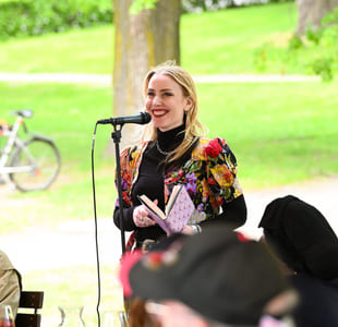 Kvinne i fargerik blomstrete jakke snakker ved mikrofon utendørs, holder en bok. Publikum er synlige i en grønn park med sykkel.