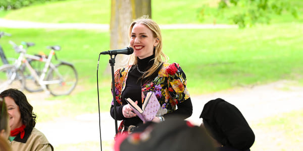 Kvinne i fargerik blomstrete jakke snakker ved mikrofon utendørs, holder en bok. Publikum er synlige i en grønn park med sykkel.