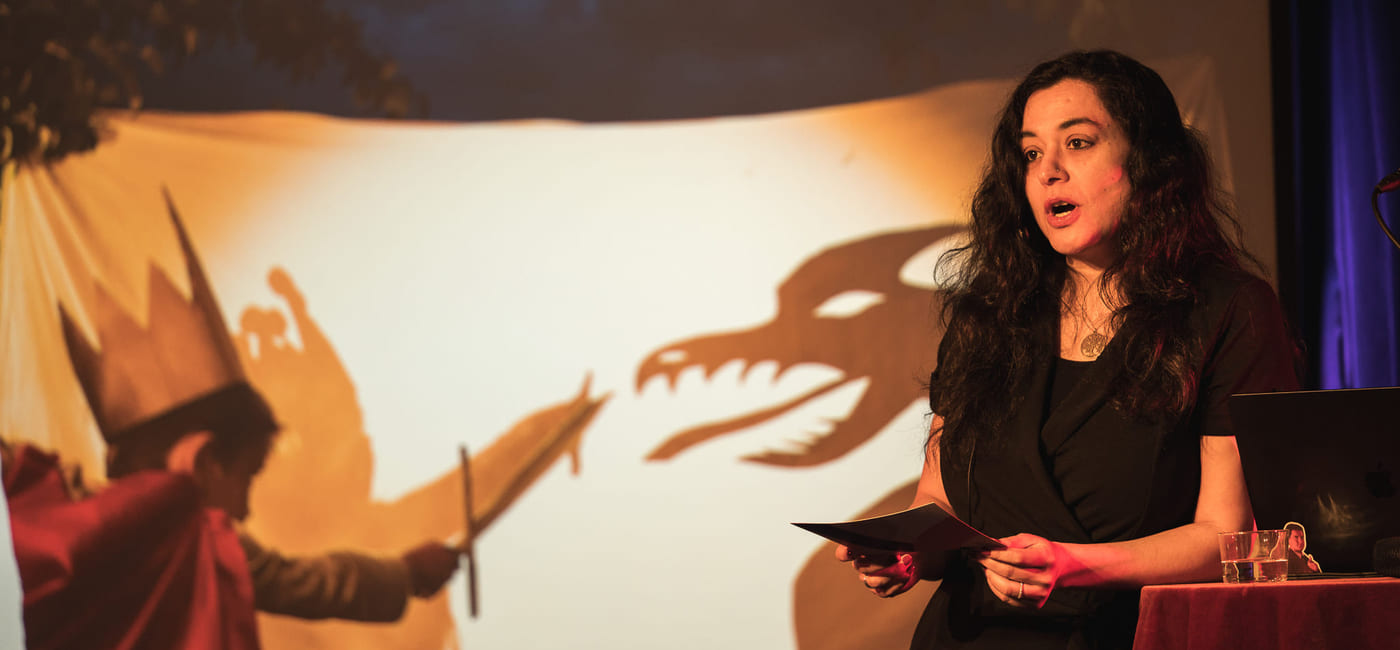 Kvinne opptrer på scenen i et intimt lokale, synlige publikumssilhuetter, varm belysning, mikrofonstativ og lampe i nærheten.