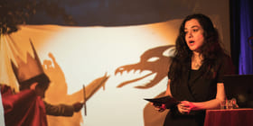 Kvinne opptrer på scenen i et intimt lokale, synlige publikumssilhuetter, varm belysning, mikrofonstativ og lampe i nærheten.