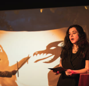 Kvinne opptrer på scenen i et intimt lokale, synlige publikumssilhuetter, varm belysning, mikrofonstativ og lampe i nærheten.