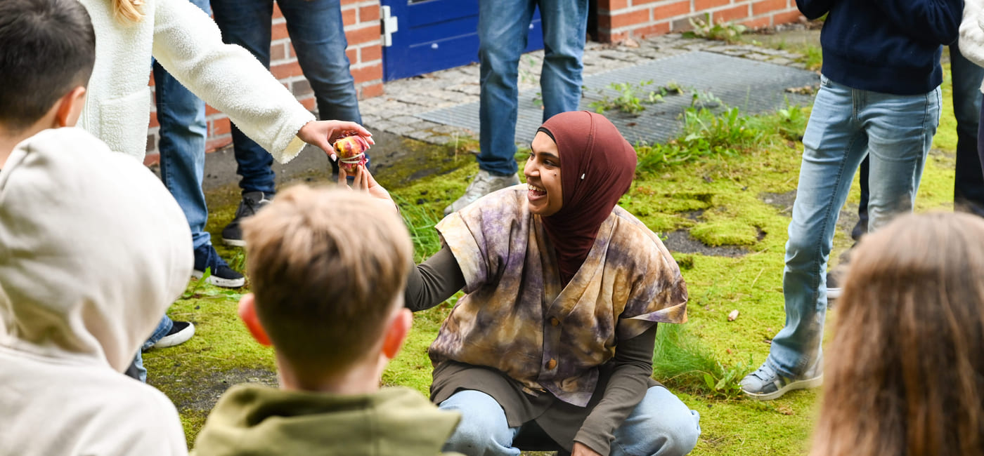 Kvinne i hijab samhandler med barn utendørs og deler ut tøybiter fra en kjele mens hun er omgitt av voksne og barn.