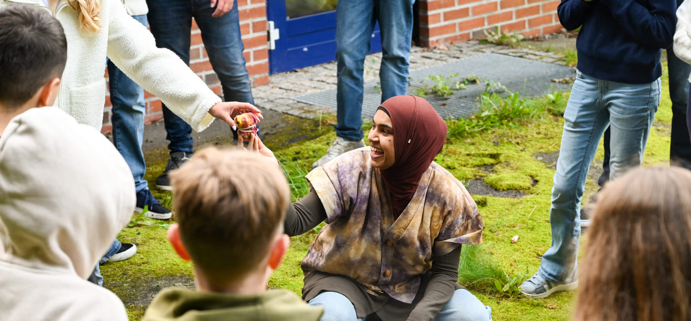 Kvinne i hijab samhandler med barn utendørs og deler ut tøybiter fra en kjele mens hun er omgitt av voksne og barn.