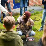 Kvinne i hijab samhandler med barn utendørs og deler ut tøybiter fra en kjele mens hun er omgitt av voksne og barn.