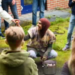 Kvinne i hijab samhandler med barn utendørs og deler ut tøybiter fra en kjele mens hun er omgitt av voksne og barn.