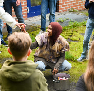 Kvinne i hijab samhandler med barn utendørs og deler ut tøybiter fra en kjele mens hun er omgitt av voksne og barn.