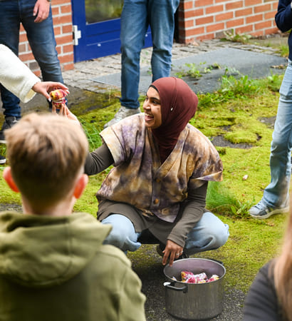 Kvinne i hijab samhandler med barn utendørs og deler ut tøybiter fra en kjele mens hun er omgitt av voksne og barn.