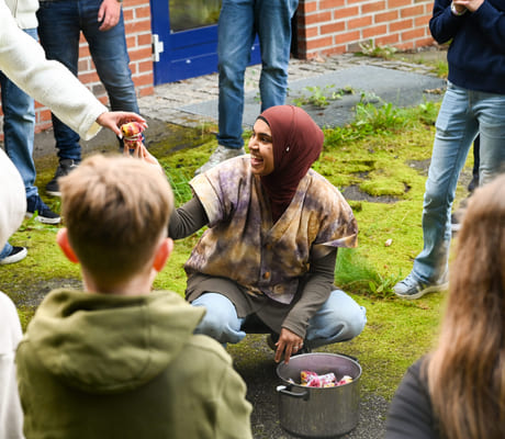 Kvinne i hijab samhandler med barn utendørs og deler ut tøybiter fra en kjele mens hun er omgitt av voksne og barn.