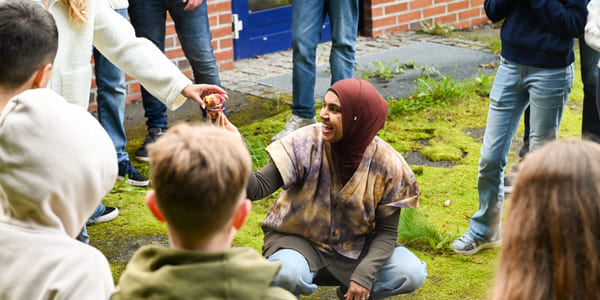 Kvinne i hijab samhandler med barn utendørs og deler ut tøybiter fra en kjele mens hun er omgitt av voksne og barn.
