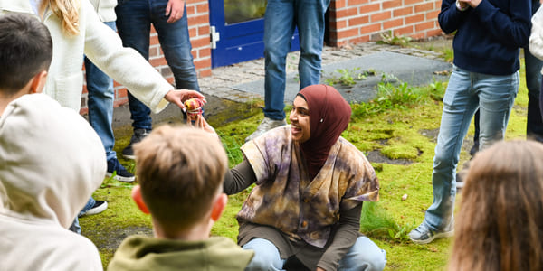 Kvinne i hijab samhandler med barn utendørs og deler ut tøybiter fra en kjele mens hun er omgitt av voksne og barn.