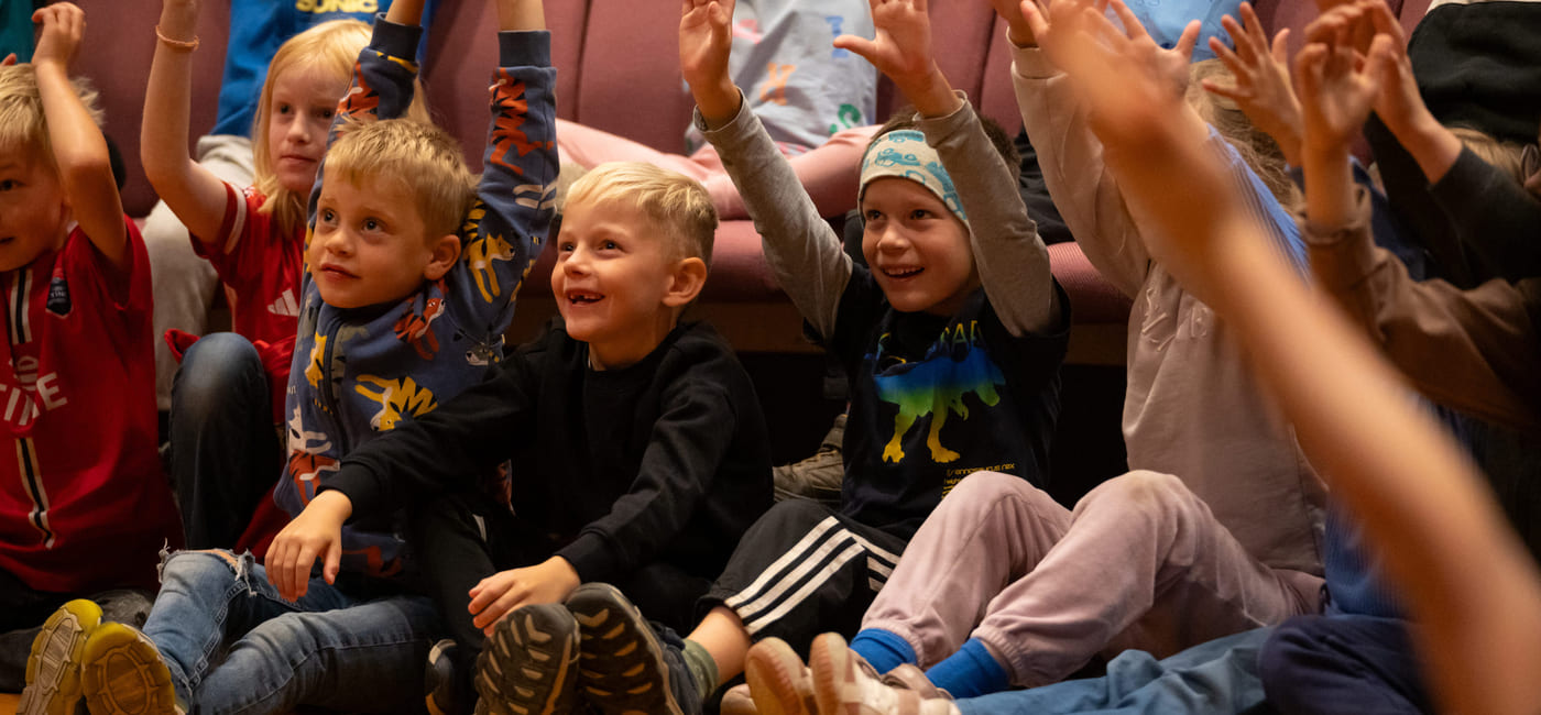 Gruppe spente barn som sitter på gulvet med løftede armer og deltar i en begivenhet eller aktivitet.