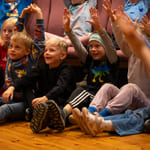 Gruppe spente barn som sitter på gulvet med løftede armer og deltar i en begivenhet eller aktivitet.