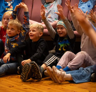 Gruppe spente barn som sitter på gulvet med løftede armer og deltar i en begivenhet eller aktivitet.