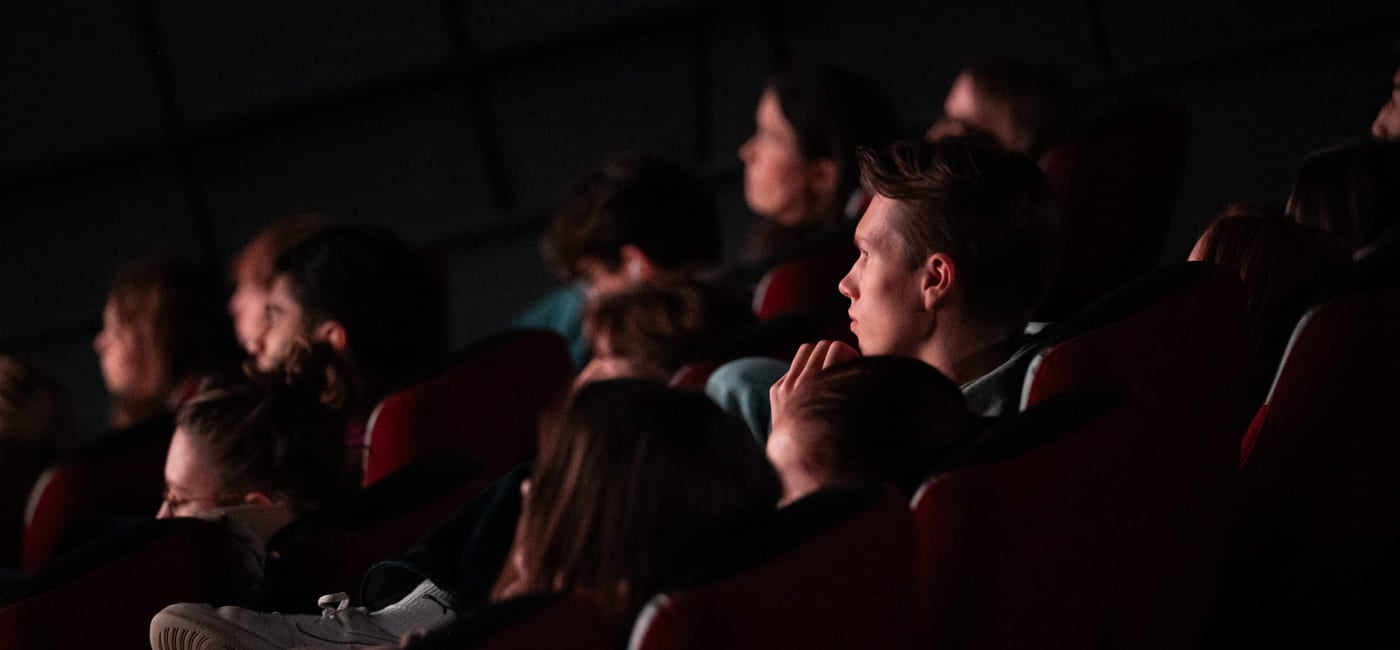 Folk sitter i et mørkt teater, fokusert på en usett forestilling. Én persons fot er synlig i forgrunnen. Dempet belysning fremhever det oppmerksomme publikummet.