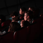 Folk sitter i et mørkt teater, fokusert på en usett forestilling. Én persons fot er synlig i forgrunnen. Dempet belysning fremhever det oppmerksomme publikummet.