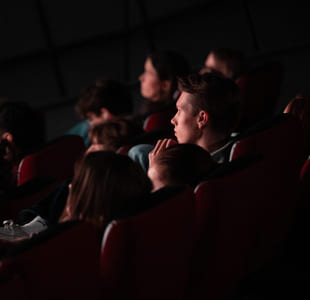 Folk sitter i et mørkt teater, fokusert på en usett forestilling. Én persons fot er synlig i forgrunnen. Dempet belysning fremhever det oppmerksomme publikummet.