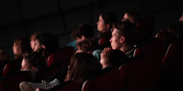 Folk sitter i et mørkt teater, fokusert på en usett forestilling. Én persons fot er synlig i forgrunnen. Dempet belysning fremhever det oppmerksomme publikummet.