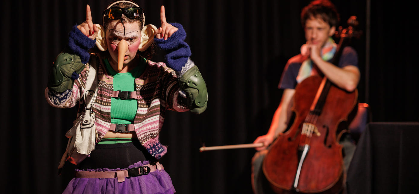 To utøvere på scenen: fargerikt kledd person som gestikulerer, og cellist som spiller et instrument i bakgrunnen. Teatralsk belysning.