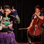 To utøvere på scenen: fargerikt kledd person som gestikulerer, og cellist som spiller et instrument i bakgrunnen. Teatralsk belysning.